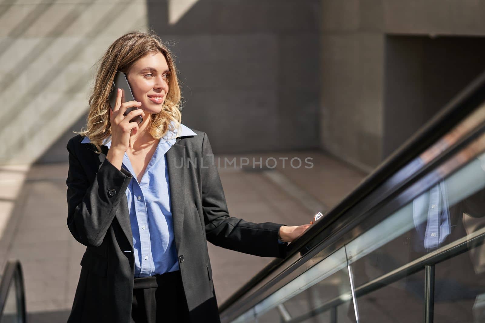 Smiling successful business woman talking on mobile phone while going up on escalator, walking in city.