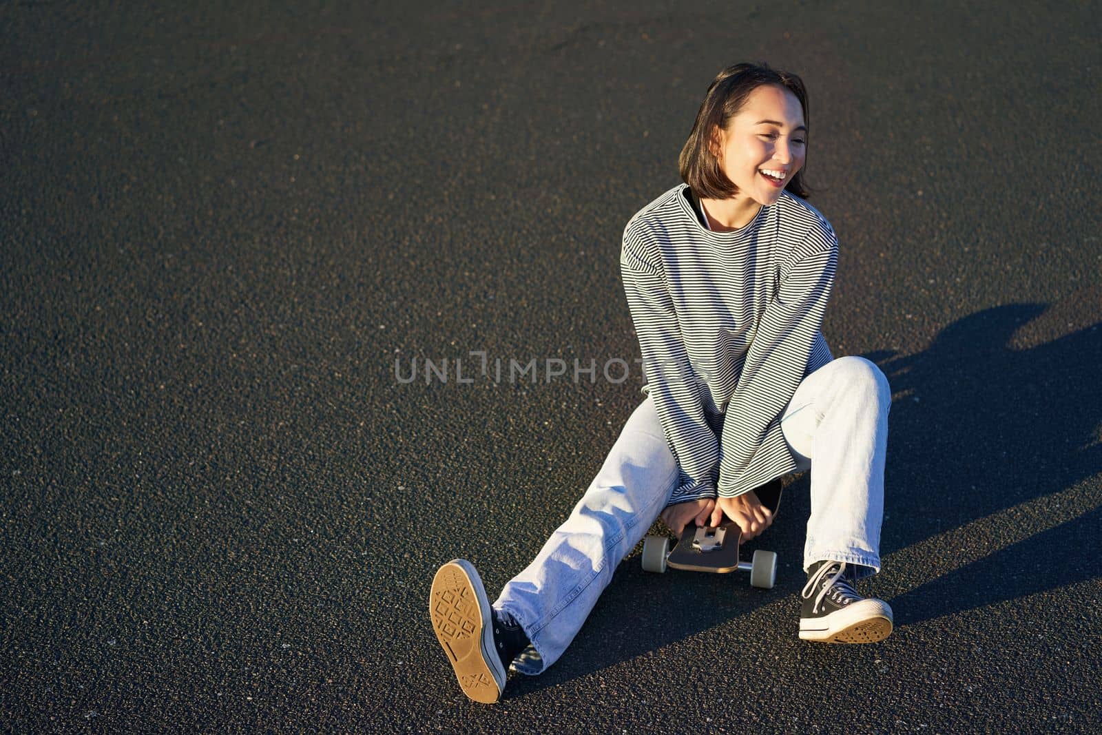 Happy beautiful korean teen girl sits on her skateboard, cruising on longboard, wearing casual clothes.