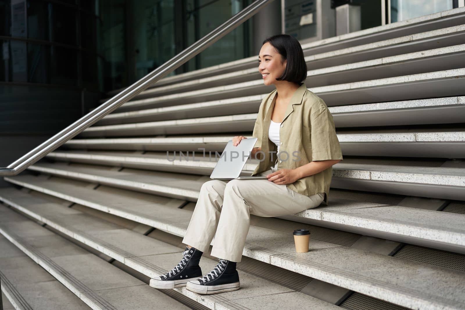 Young smiling asian girl with laptop, sits on street of city with coffee. Young woman does homework on computer while sitting on street stairs, digital nomad works remote by Benzoix