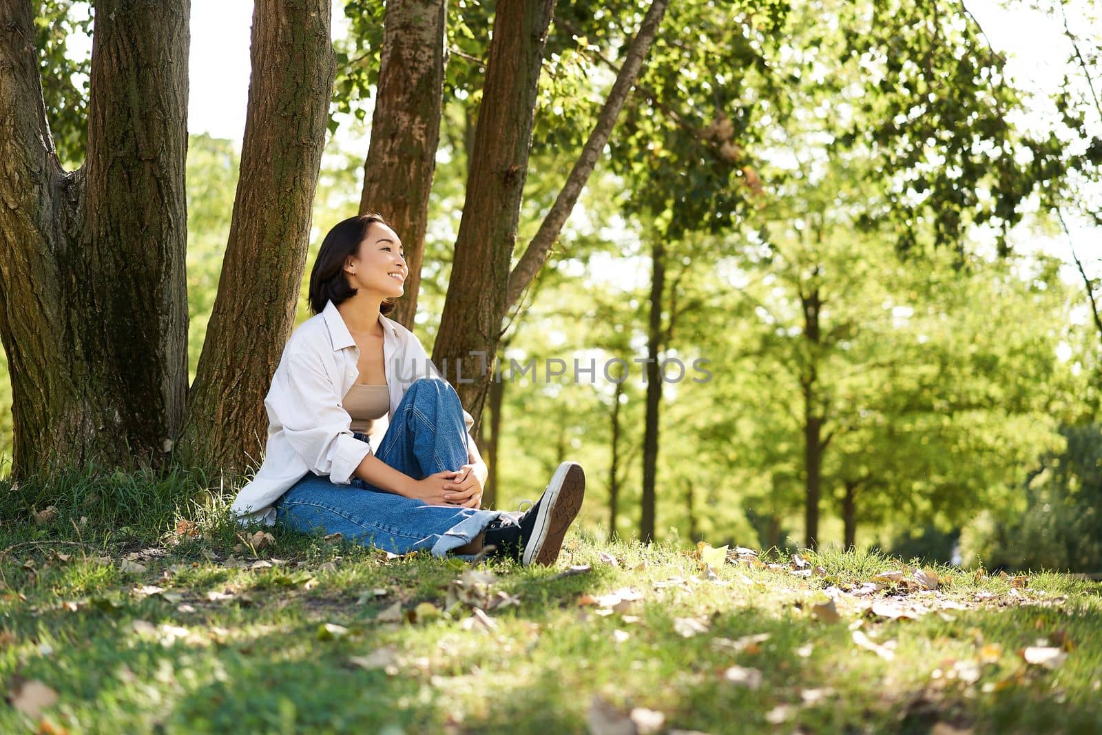 Portrait of asian girl relaxing, leaning on tree and resting in park under shade, smiling and enjoying the walk outdoors by Benzoix