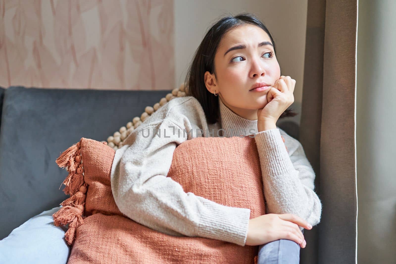 Women and wellbeing concept. Portrait of thoughtful asian woman sitting on sofa with pillow, looking aside with thinking concentrated face.