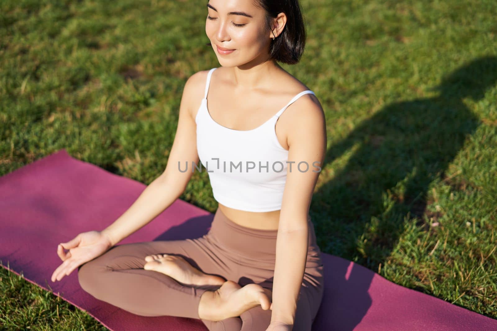 Portrait of smiling asian woman meditating, doing yoga on fresh air, relaxing on rubber mat, exercising in park, breathing air, being calm by Benzoix