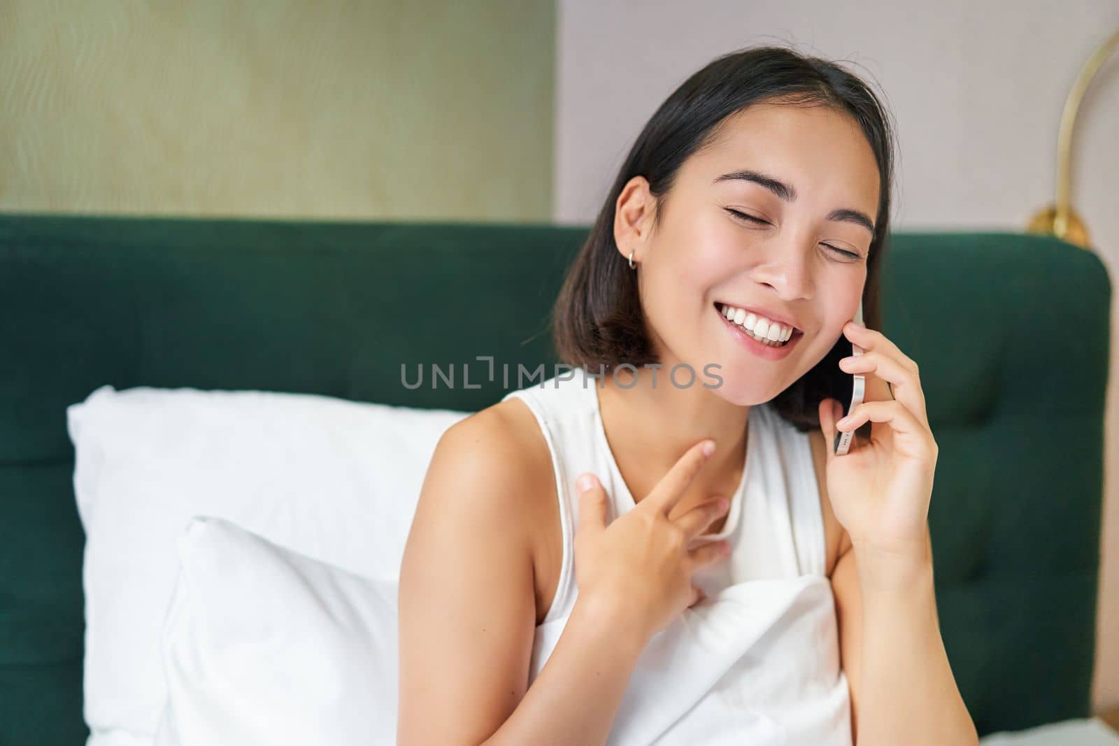 Close up portrait of cute asian girl in bed, talking on mobile phone with happy smiling face. Woman waking up and making a telephone call.