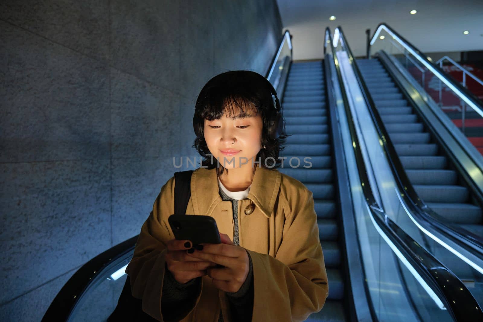 Cute young woman going down the escalator to the tube, using subway metro to commute to work or university, standing with smartphone.