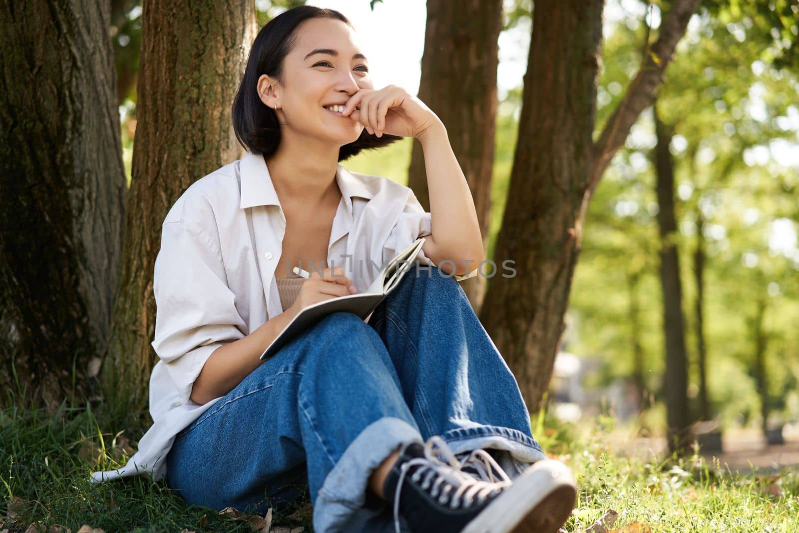 Portrait of young asian woman writing in her notebook, expressing her thoughts on paper in diary, smiling and sitting under tree on summer day in park by Benzoix