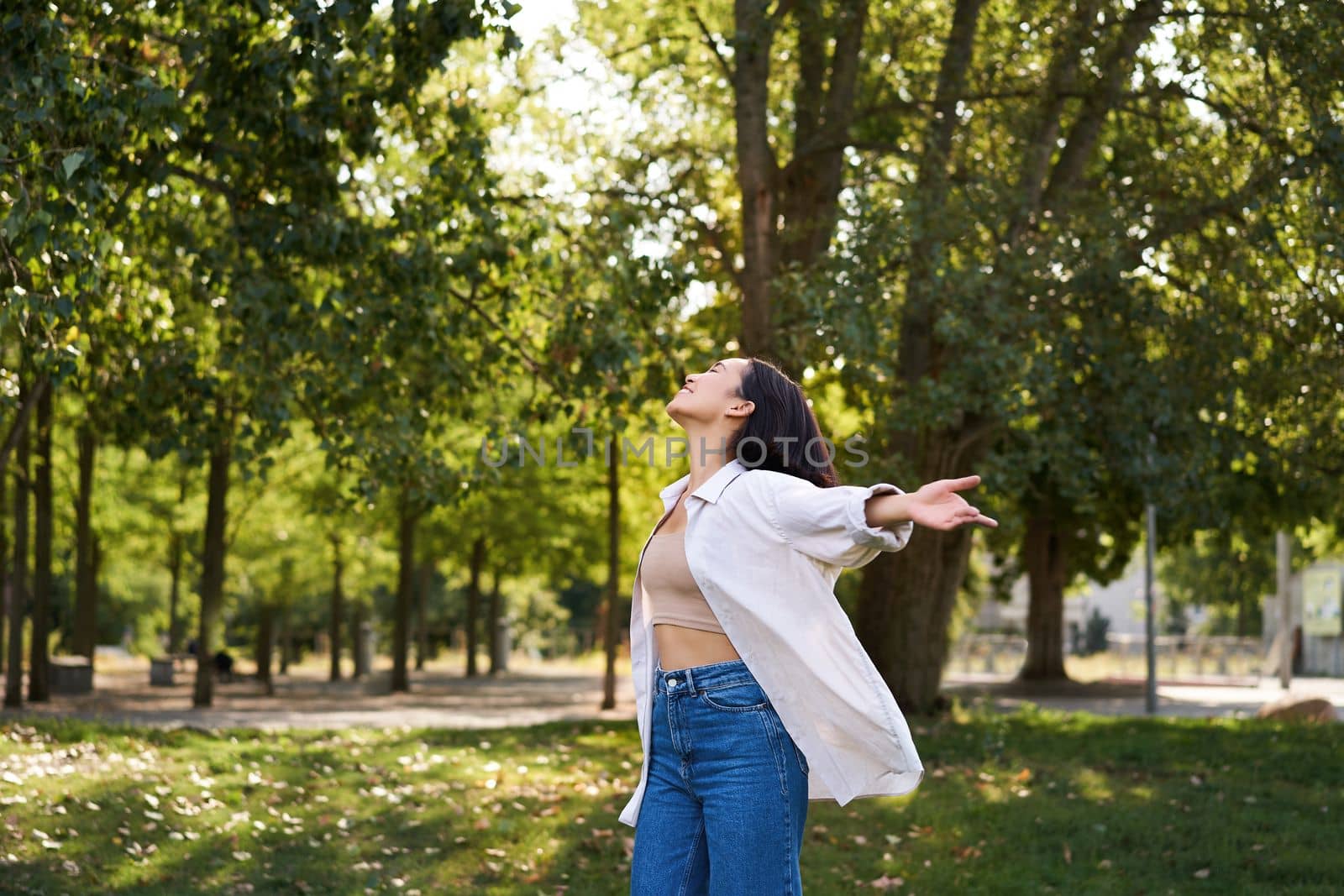 Carefree asian girl dancing, feeling happiness and joy, enjoying the sun on summer day, walking in park with green trees.