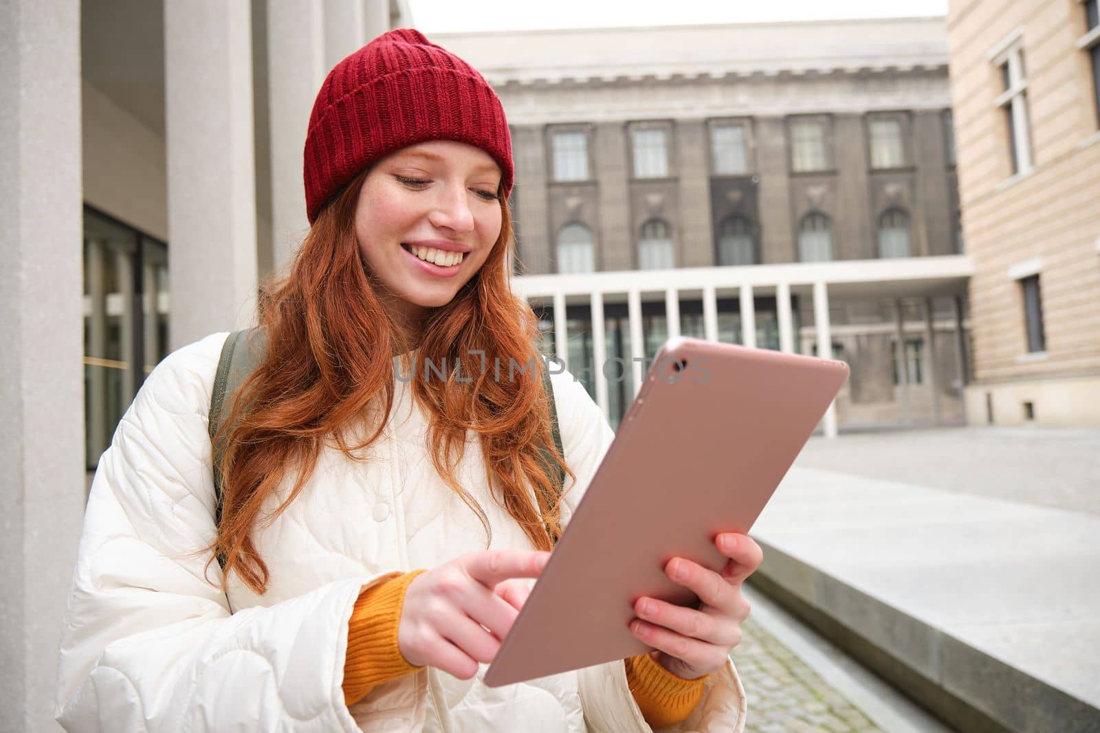 Happy redhead girl in red hat, walks around city with digital tablet, connects to public internet wifi and looks for route, looks at map on her gadget.