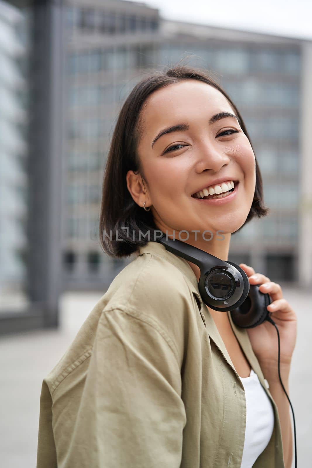 Vertical shot of beautiful asian woman posing with headphones around neck, smiling and laughing, standing on street in daylight.