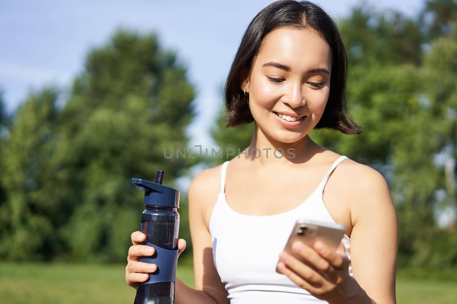 Smiling fitness girl drinks water, checks her app on smartphone and looks happy, stays hydrated on fresh air, sunny day in park by Benzoix