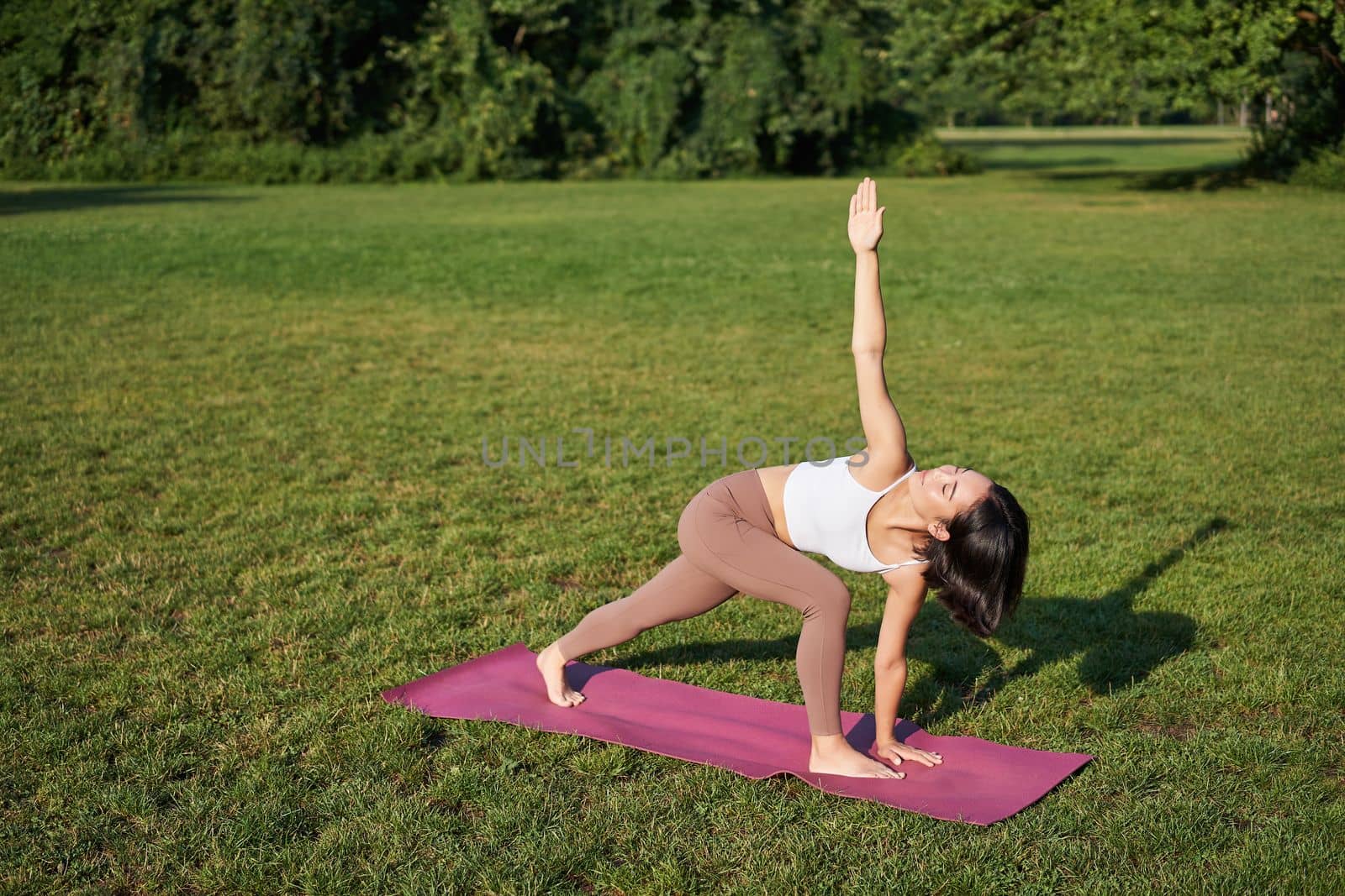 Portrait of young asian woman stretching, doing yoga on rubber mat, exercising in park, mindful training on fresh air.