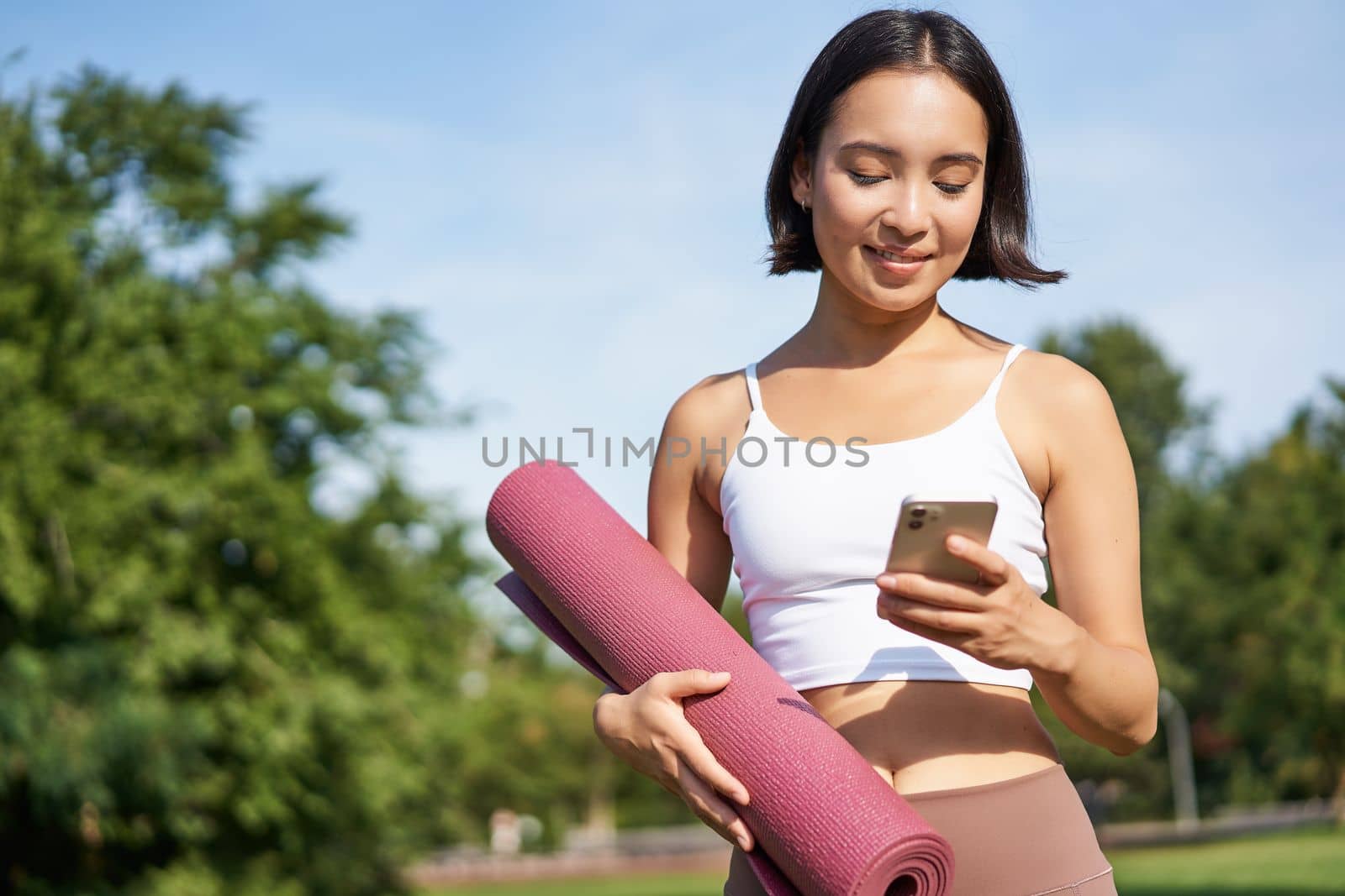 Portrait of young asian woman checking her phone during workout, walking in park with rubber mat, going to the gym, holding smartphone.