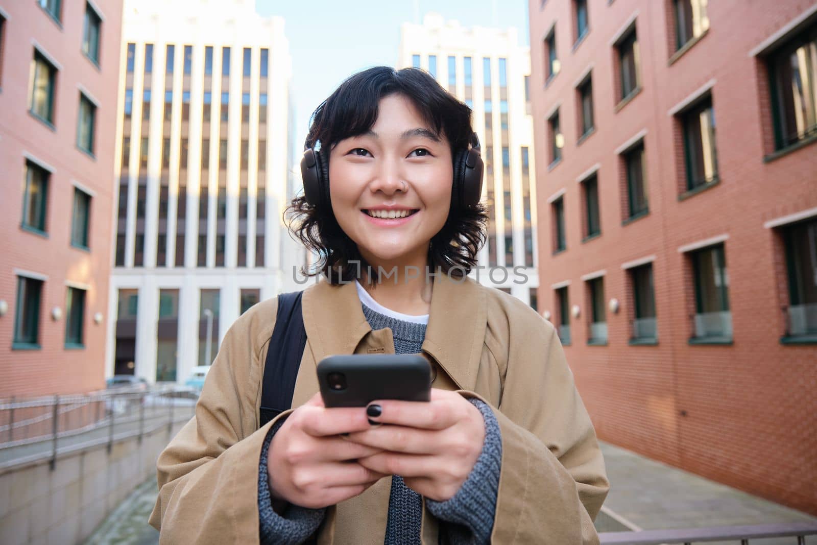 Portrait of cute girl tourist, korean woman in headphones, looks at mobile phone, uses smartphone app, map or text messages, listens music in headphones by Benzoix