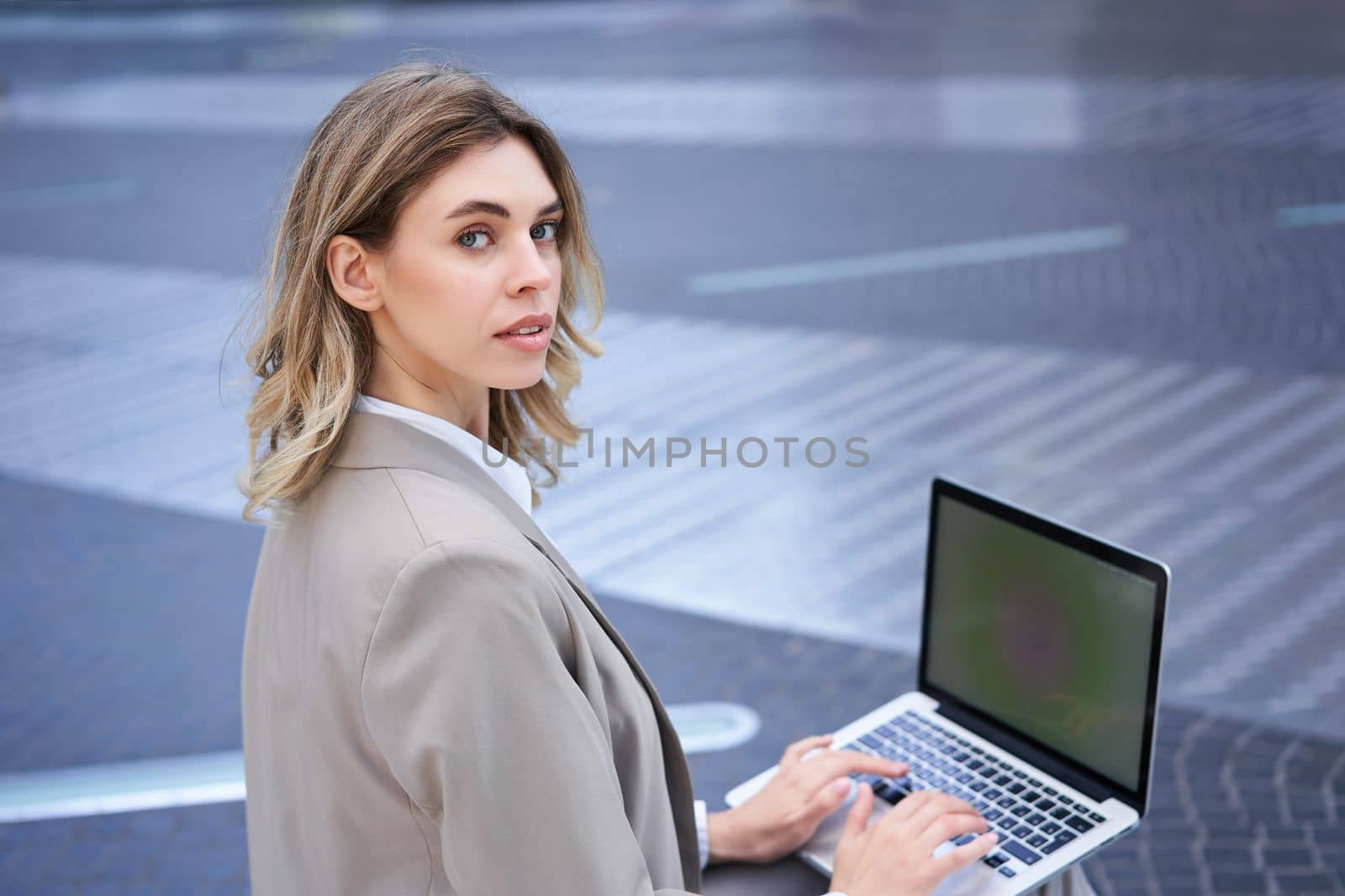 Young businesswoman using laptop while sitting outdoors in city centre, typing on keyboard. Girl preparing for interview, wearing suit.