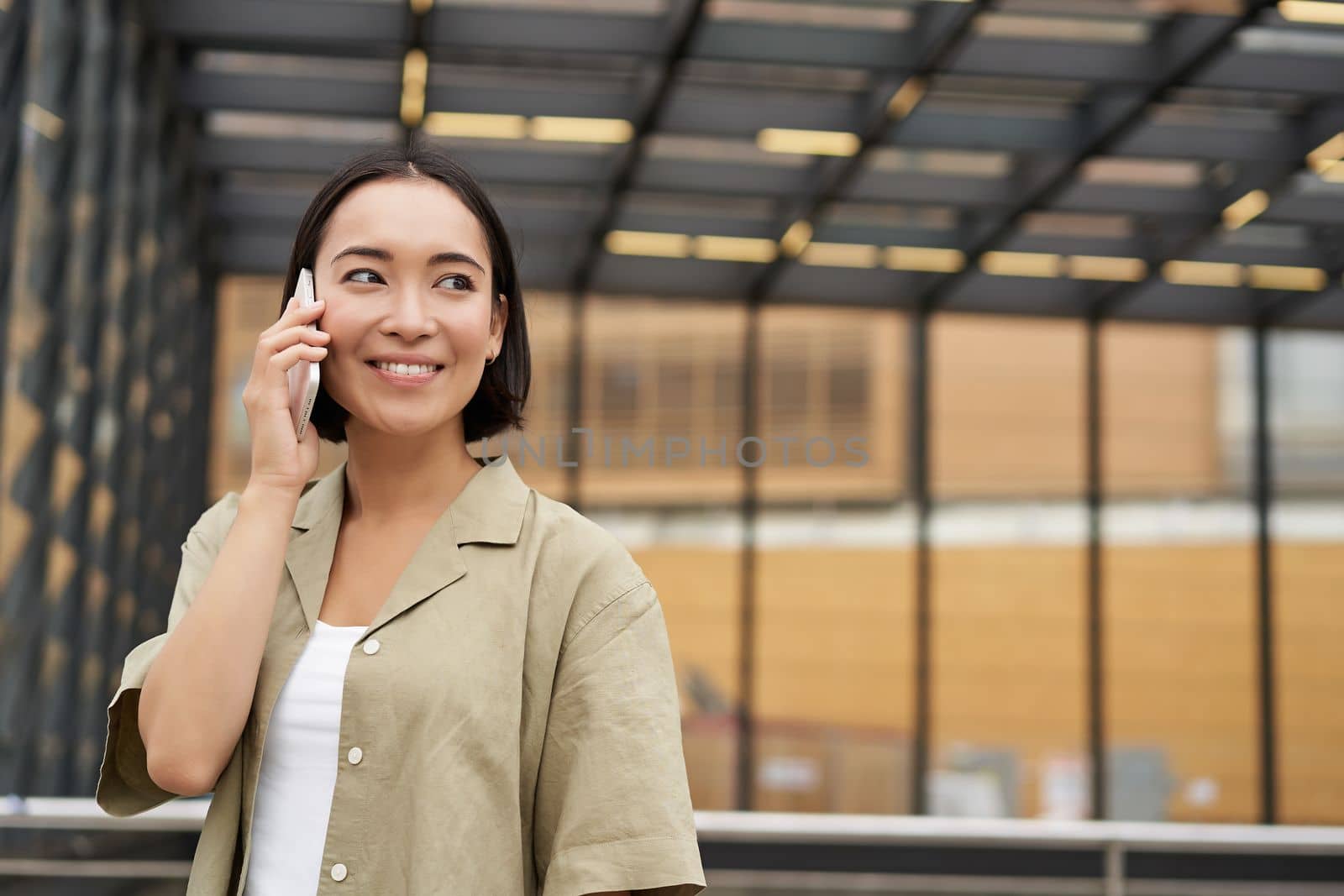 Happy asian woman talking on mobile phone, standing on bus stop in city. Outdoor shot by Benzoix