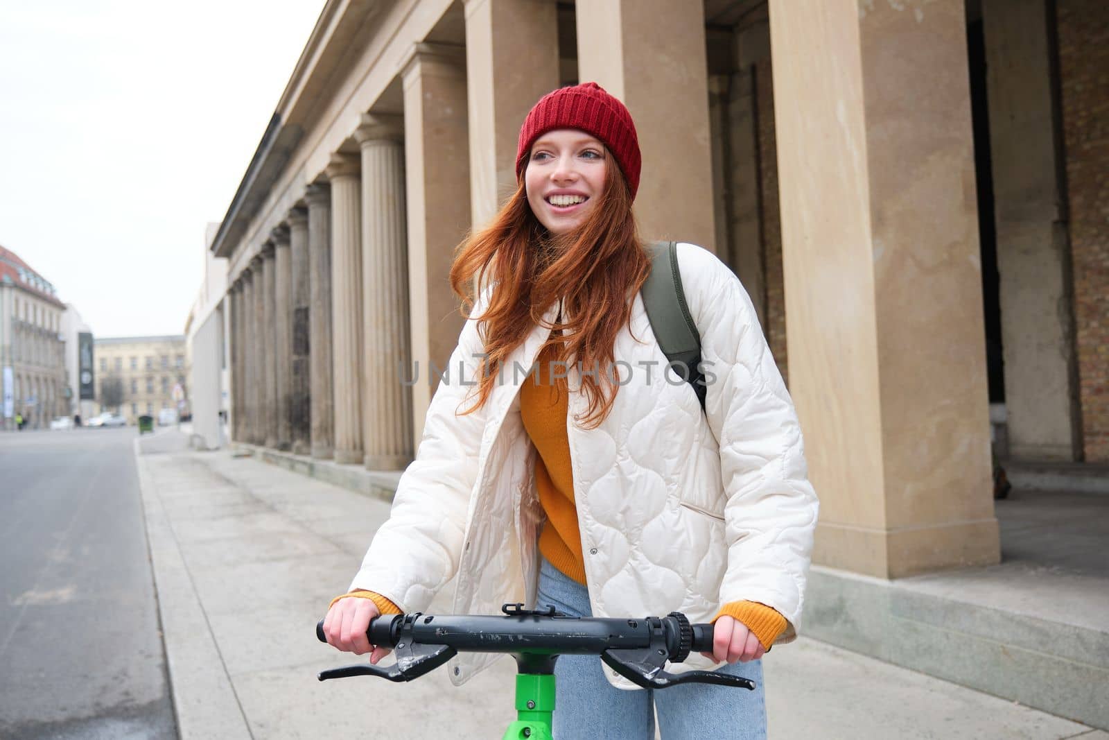 Smiling redhead european girl drives public e-scooter, tourist explores city, rides in city centre.