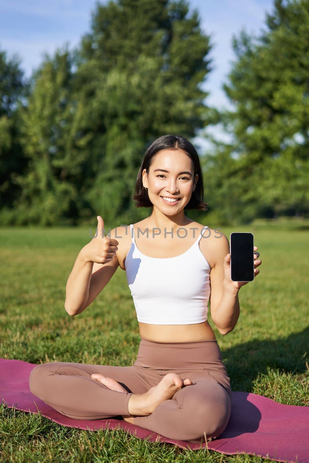 Vertical shot of asian woman shows thumbs up, recommending yoga training online, meditation app, doing exercises on fresh air in park, showing smartphone screen by Benzoix