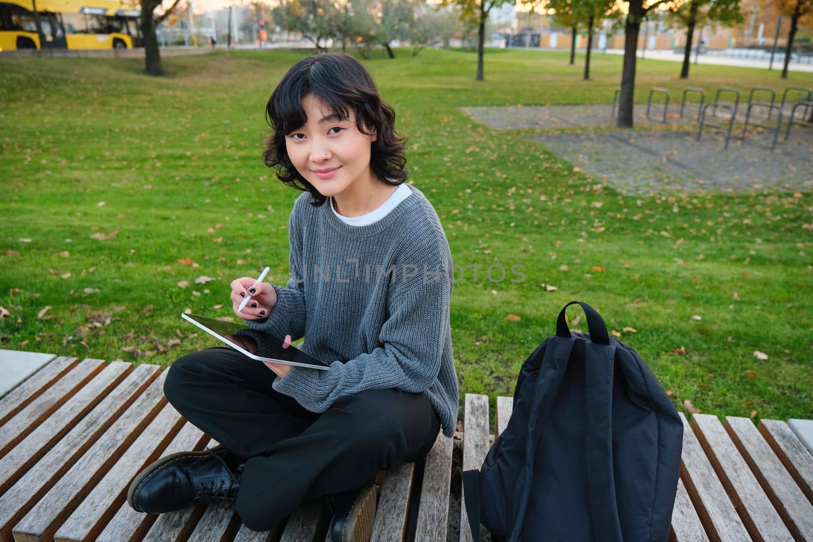 Young asian girl with graphic pencil and tablet, sits in park on bench, draws scatches, does her homework outdoors.