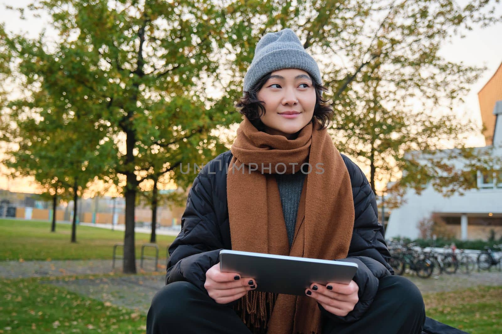 Smiling young girl, korean student sits in park in scarf and hat on chilly day, draws outdoors on digital tablet with graphic pen, makes scatches by Benzoix