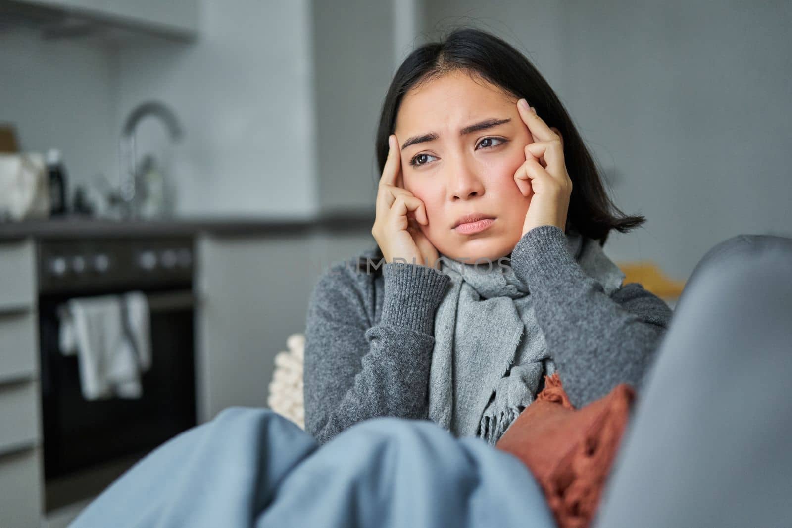 Image of sad young woman feeling sick, being unwell, sitting with scarf and frowning in her living room, catching cold and staying at home on isolation.