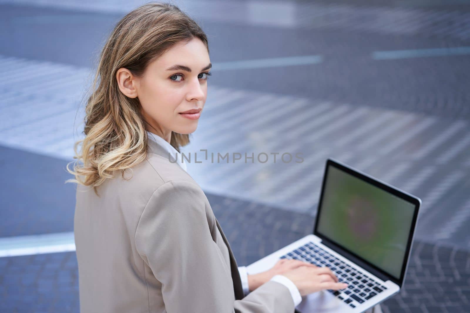 Young businesswoman using laptop while sitting outdoors in city centre, typing on keyboard. Girl preparing for interview, wearing suit.