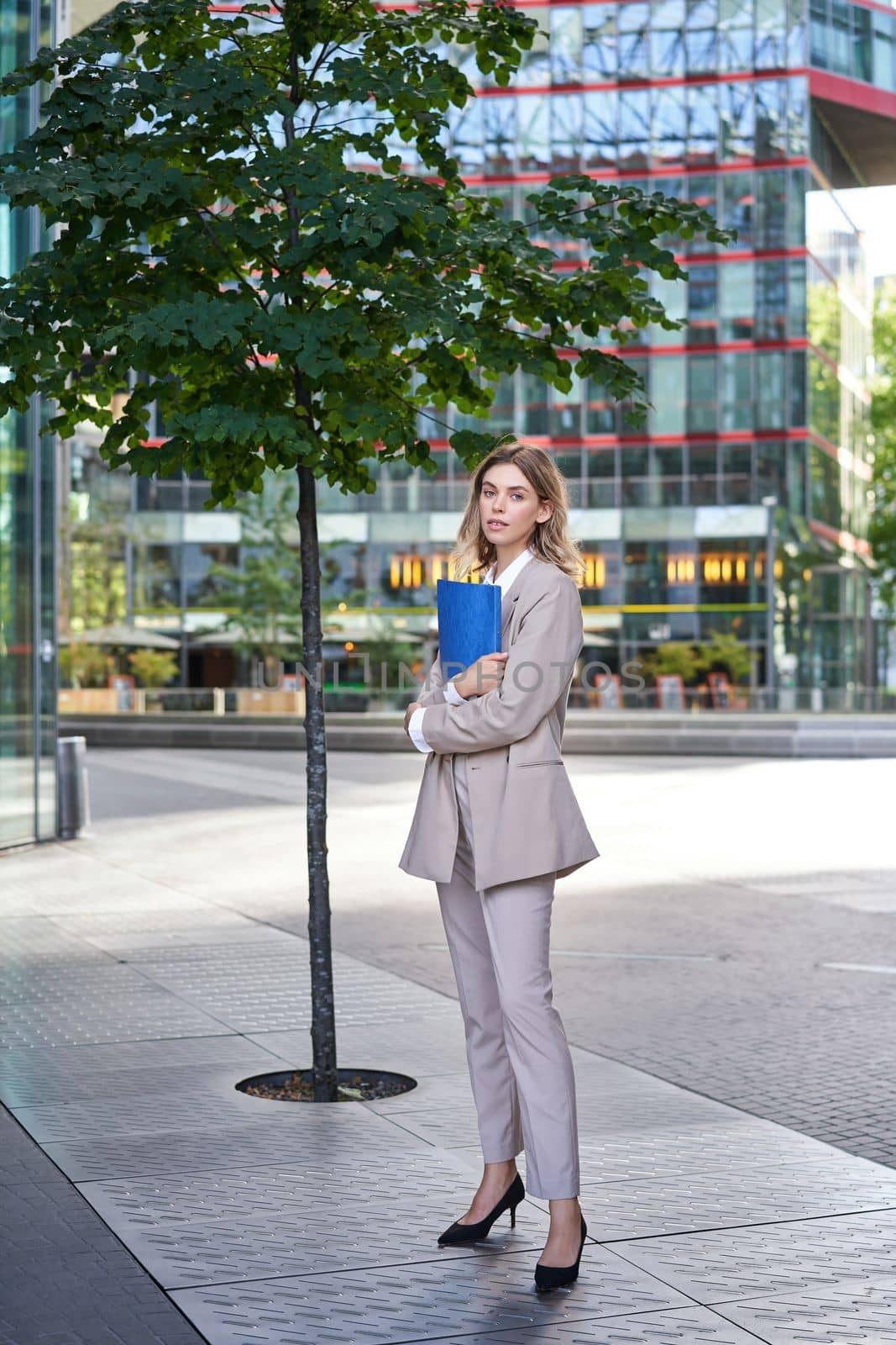 Portrait of young saleswoman in beige business suit, holding blue folder with work documents, standing outdoors on street of city center.
