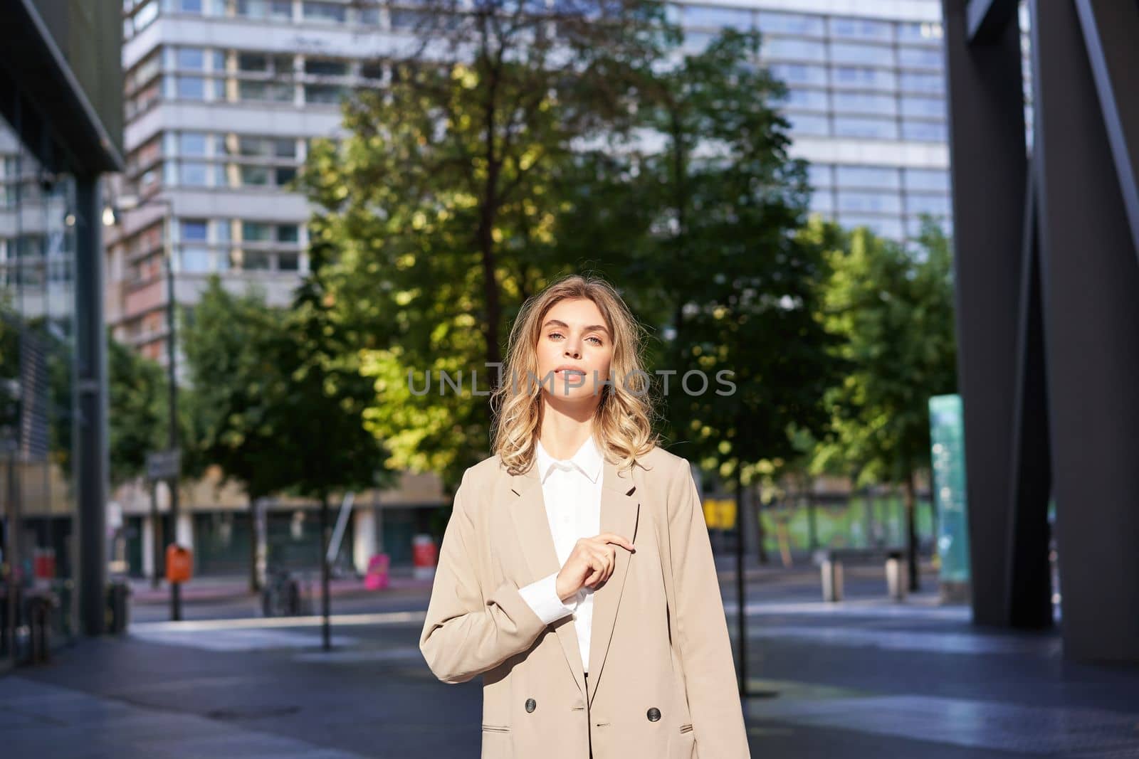 Portrait of young confident business woman in beige suit, corporate lawyer or saleswoman standing outside office building on street and smiling by Benzoix