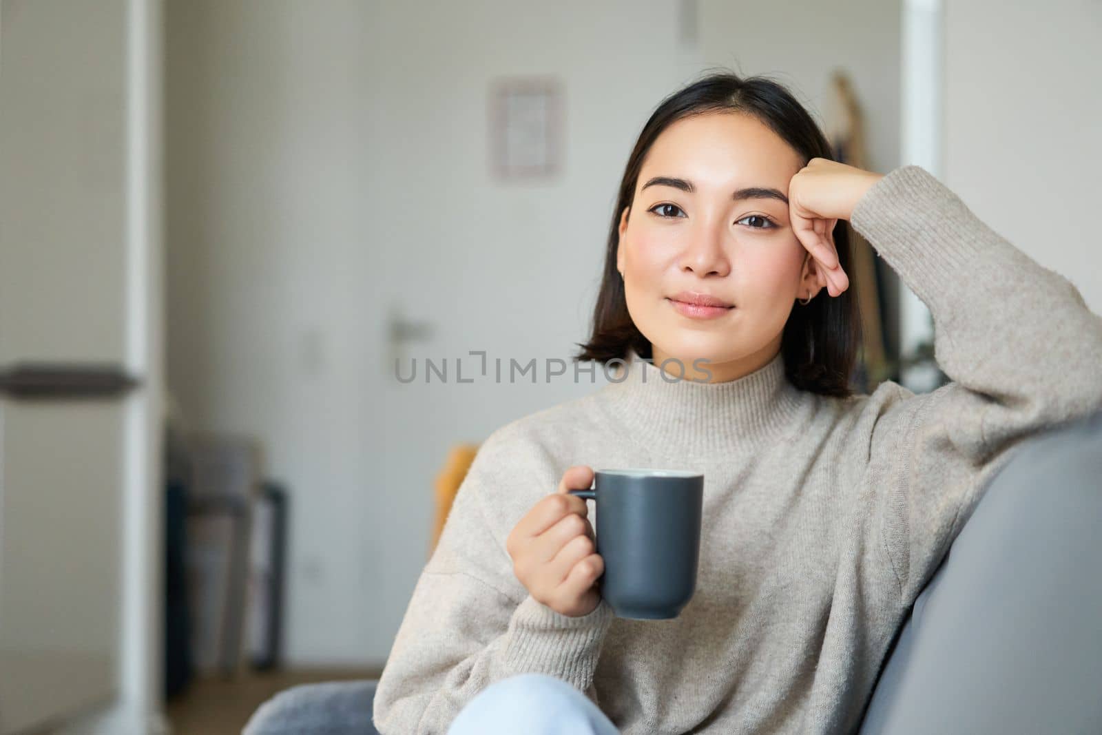 Smiling asian woman sitting on sofa with her mug, drinking coffee at home and relaxing after work, looking calm and cozy.