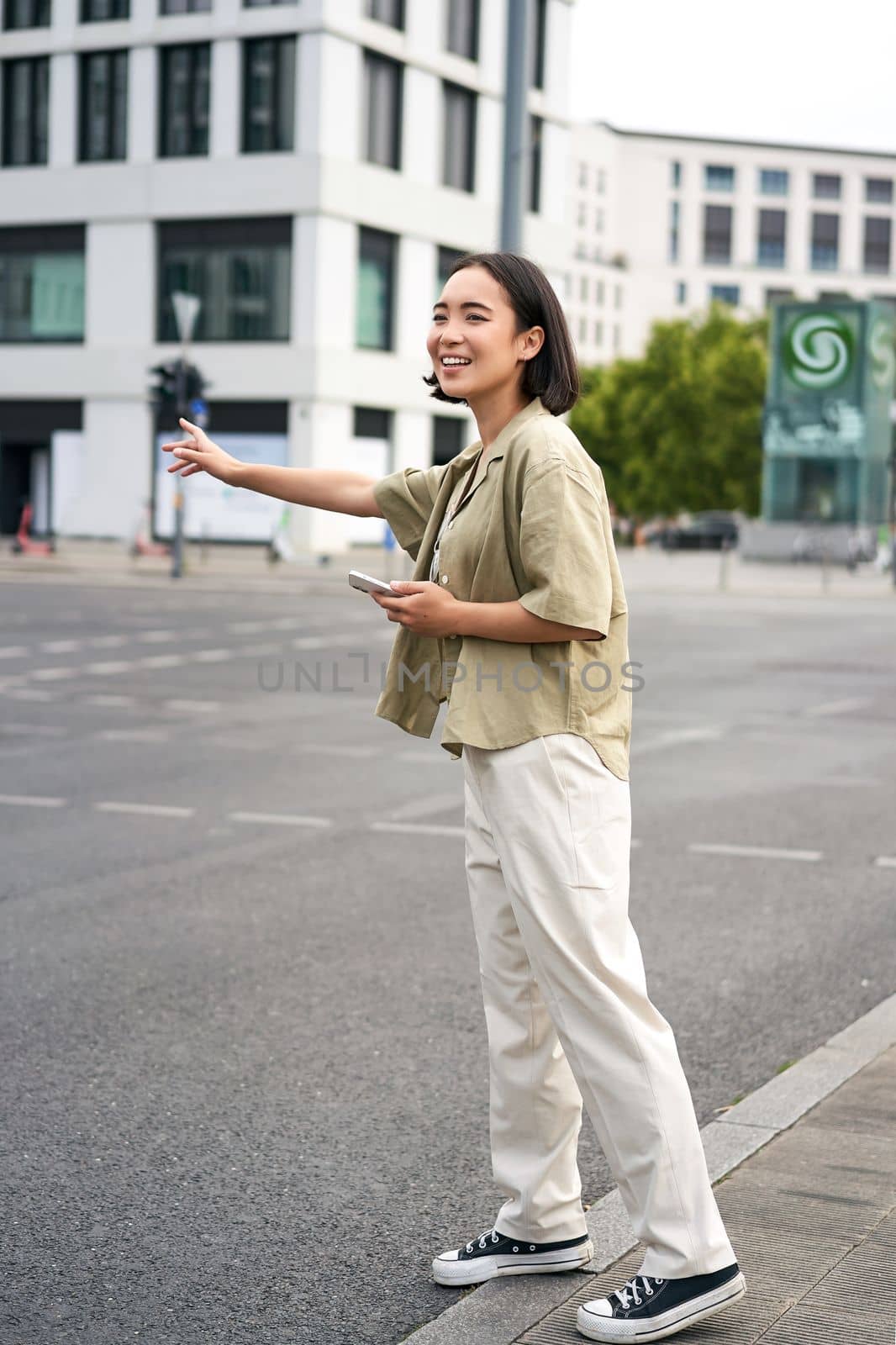 Car sharing and technology. Young asian woman waiting for taxi near road, holding smartphone, order car in application.