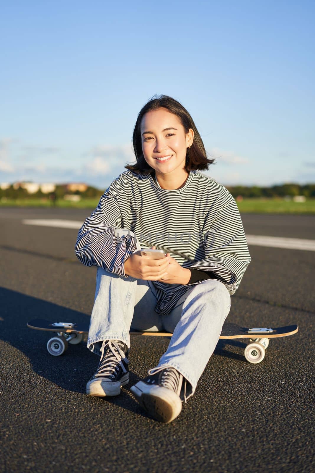 Vertical shot of asian woman sitting on skateboard on road, holding smartphone app. Skater girl skates on longboard, using mobile phone by Benzoix
