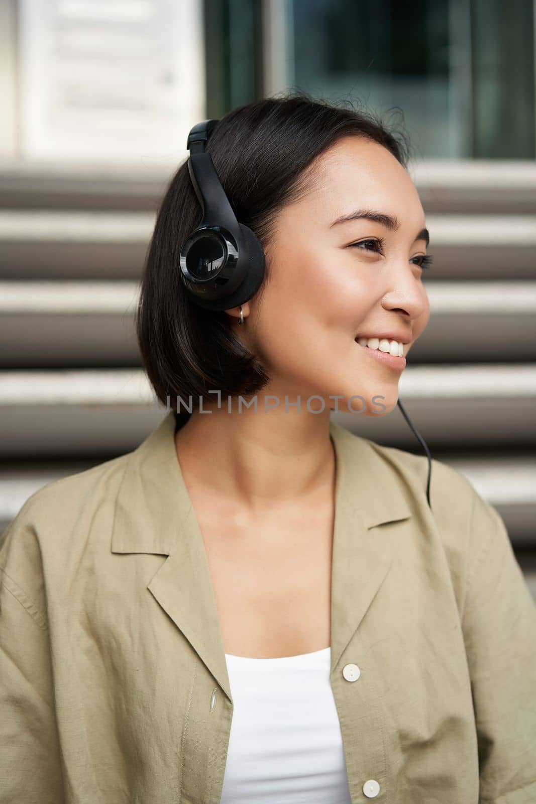 Smiling asian girl, laughing, listening music in headphones, sitting outdoors. Uni student enjoying free time.