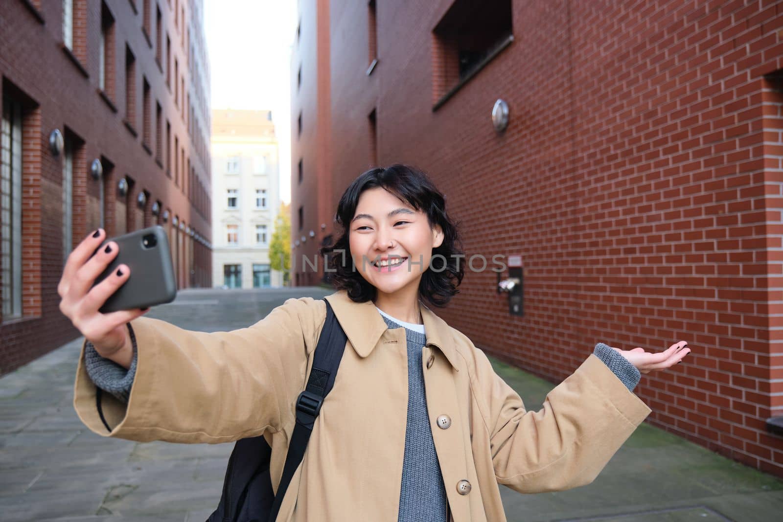 Happy korean girl, tourist takes photos on street, shows smth while records video of herself on smartphone, posing near building and smiling by Benzoix