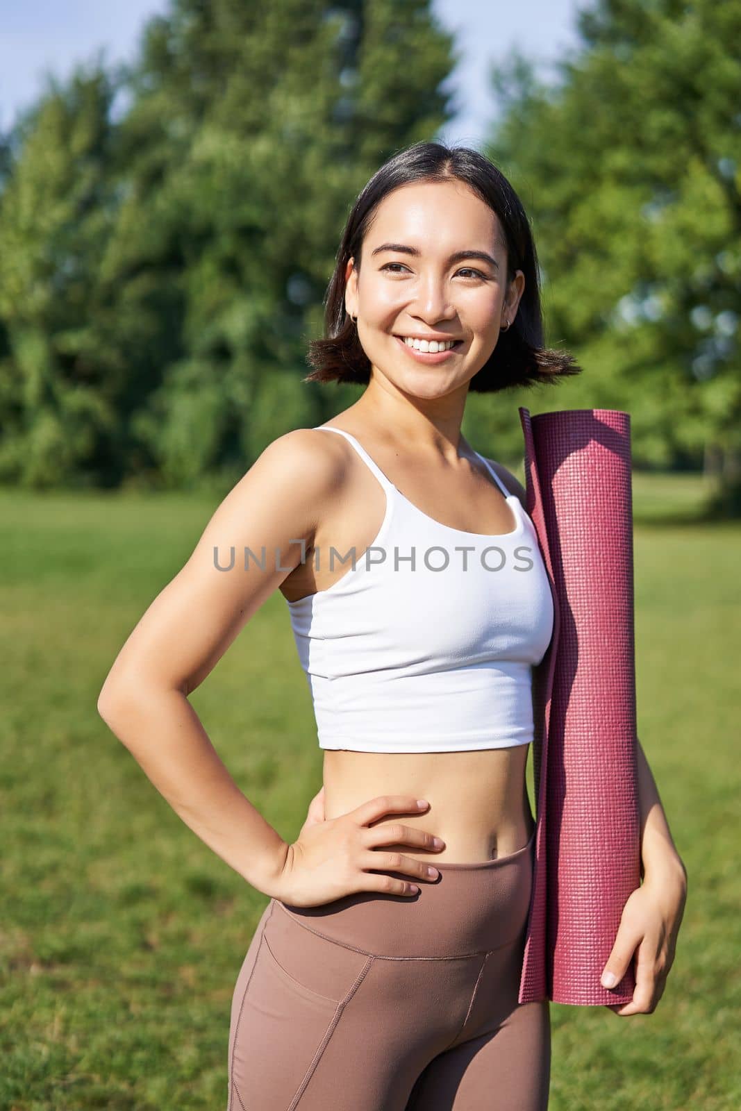 Smiling fitness girl with rubber mat, stands in park wearing uniform for workout and sport activities, does yoga outdoors on lawn.
