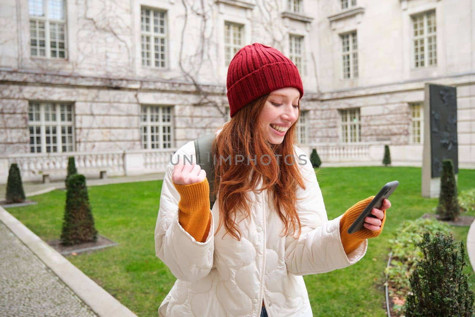 Portrait of cheerful redhead woman with smartphone, celebrating, reading amazing news and rejoicing, triumphing on street.