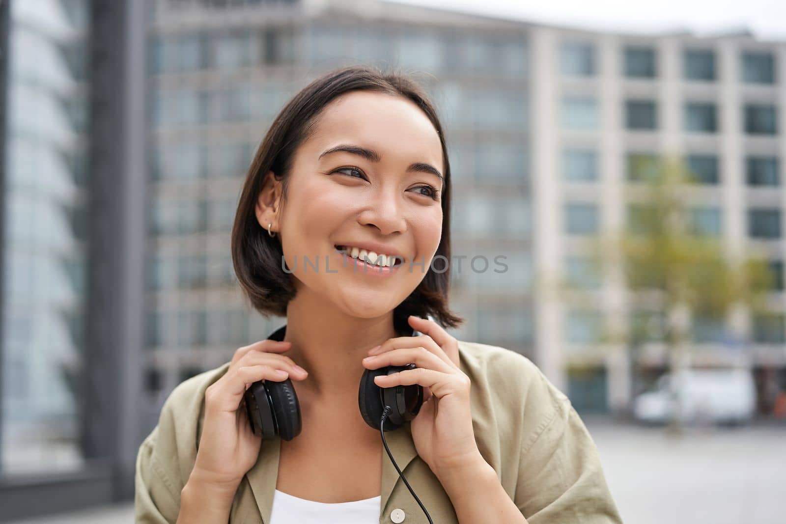 Portrait of smiling asian girl with headphones, posing in city centre, listening music.