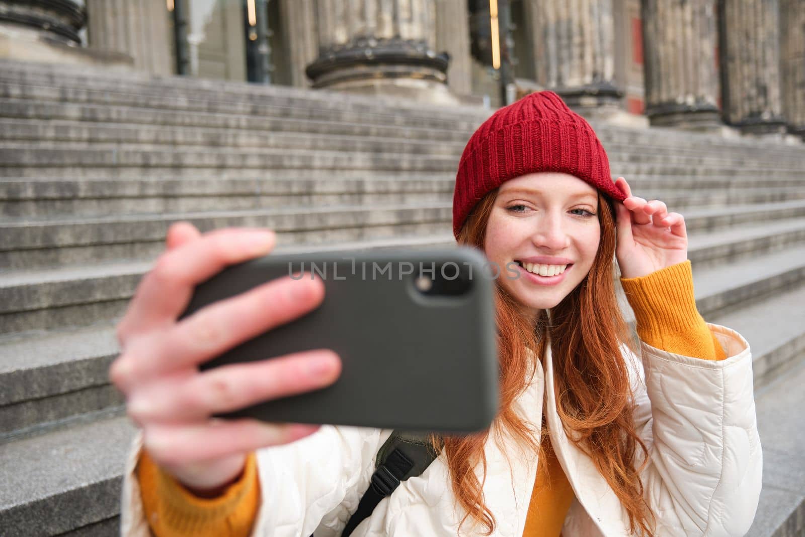 Young redhead tourist takes selfie in front of museum on stairs, holds smartphone and looks at mobile camera, makes photo of herself with phone by Benzoix