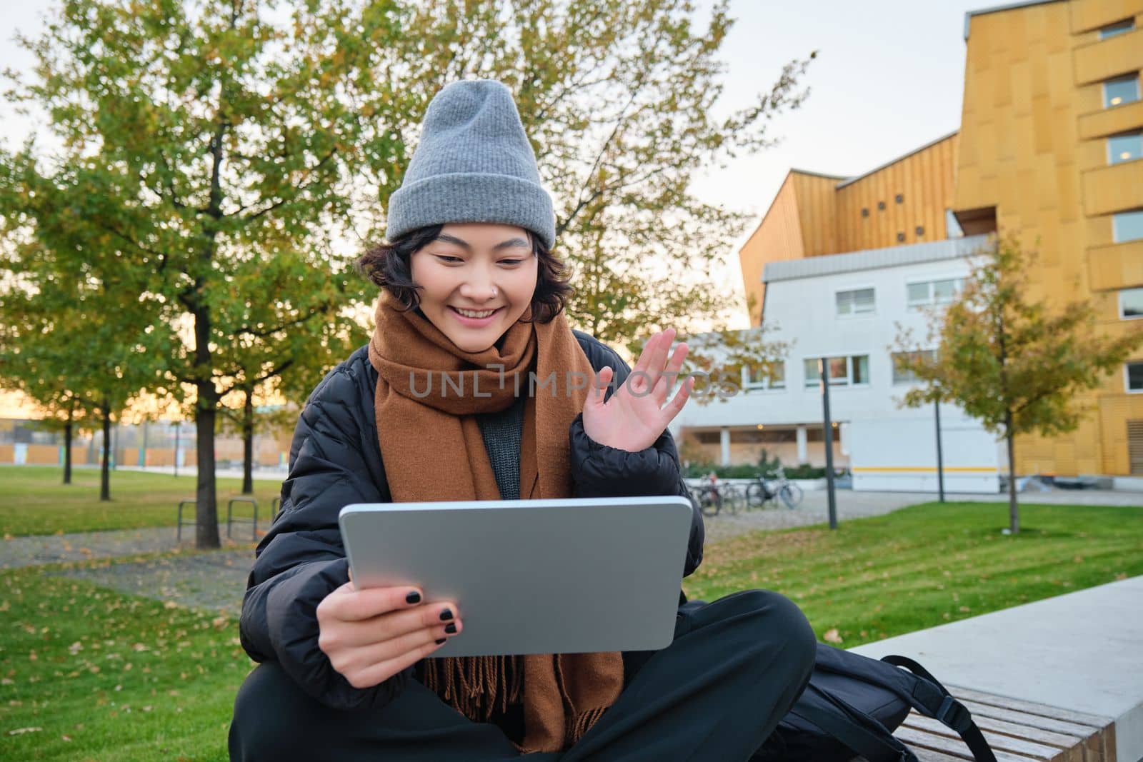 Portrait of young asian girl sits in warm clothes in park, waves hand at tablet, video chats outdoors, says hello while on call.