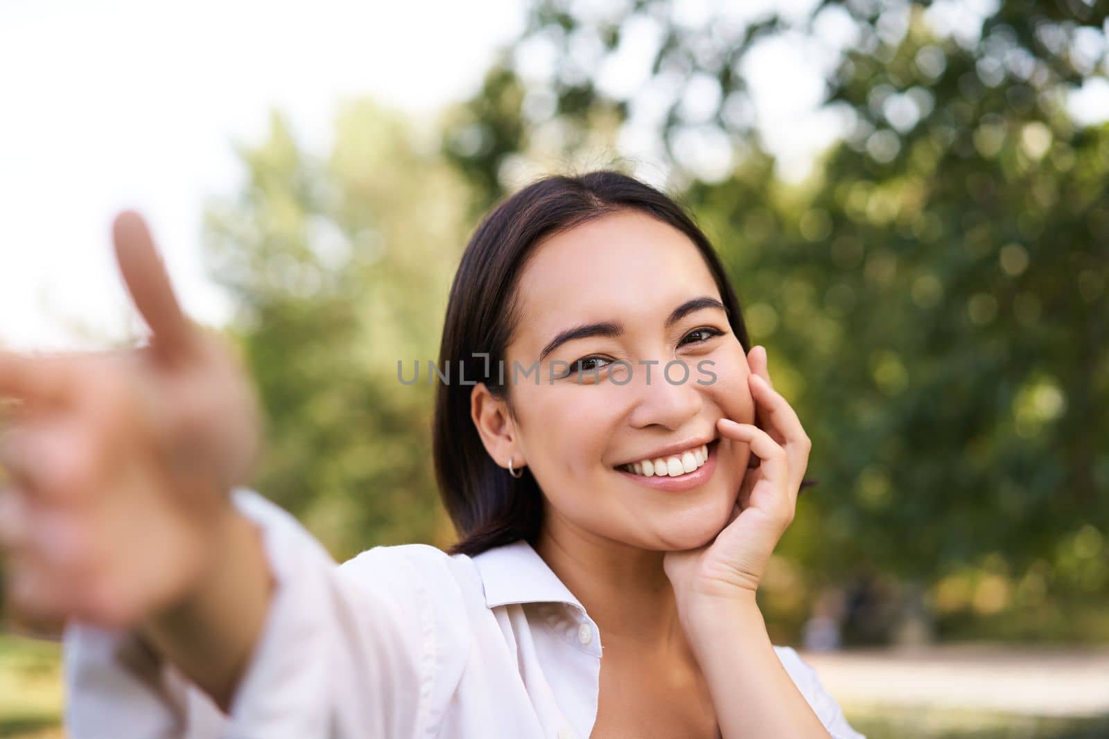 Smiling asian girl takes selfie, video chats, holds smartphone and speaks at camera, posing in park.