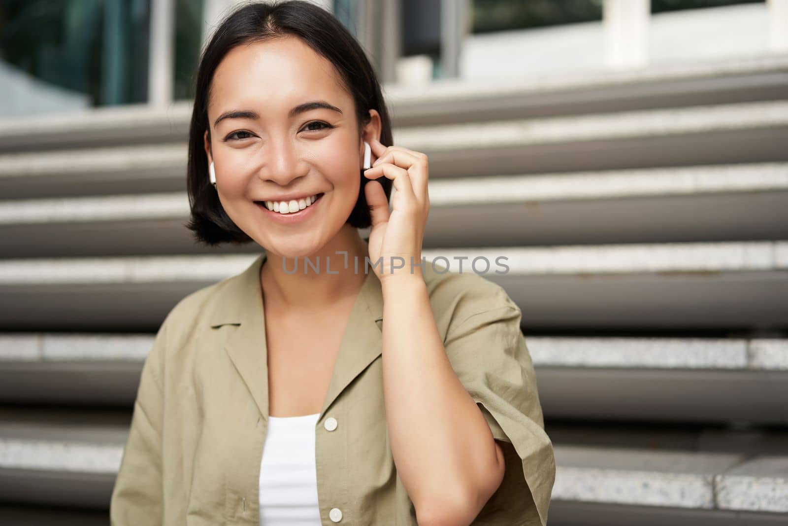 Portrait of smiling asian girl listens music, podast in wireless earphones, using headphones outdoors, sitting on street.