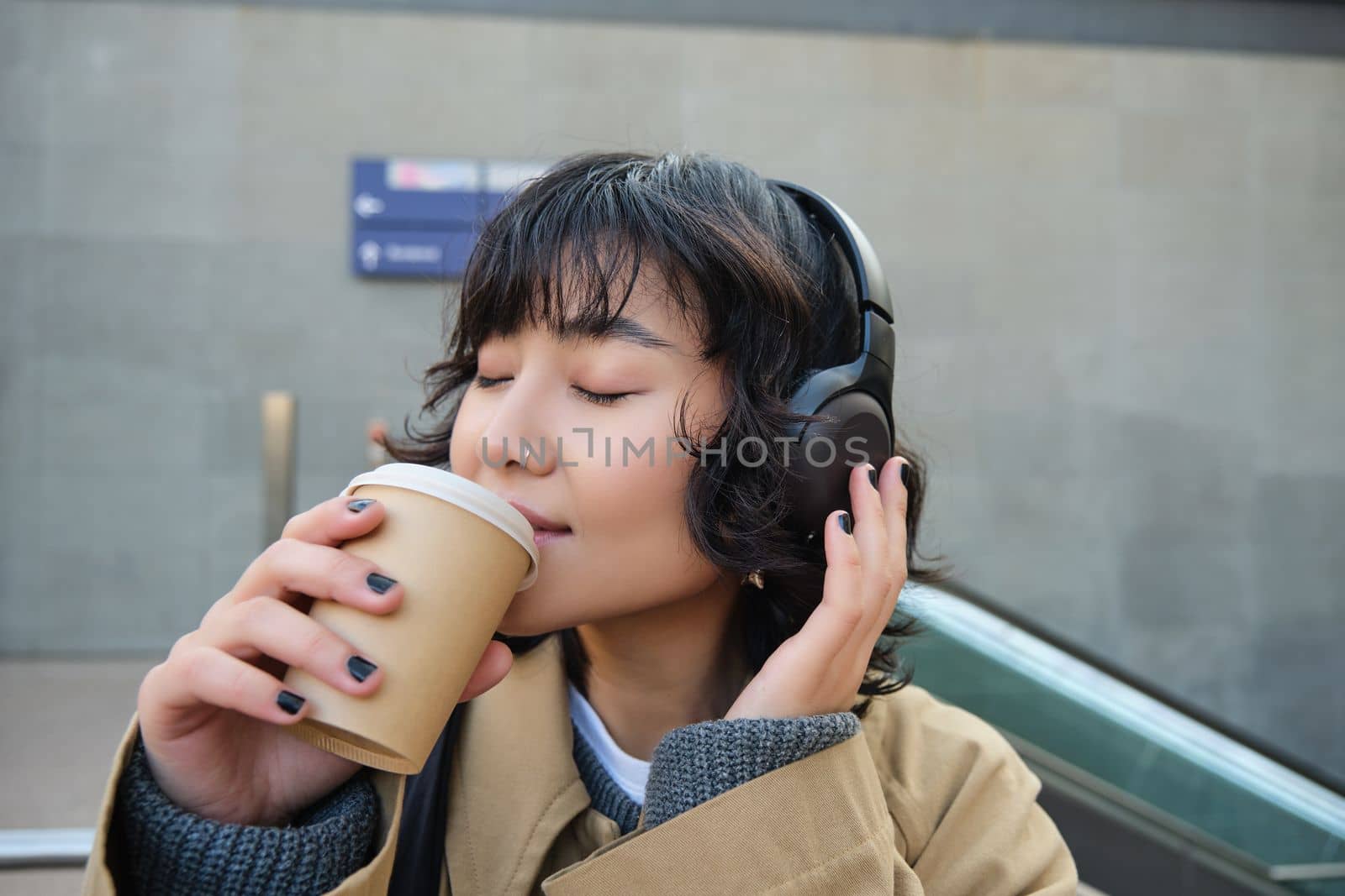 Happy brunette woman in headphones, listens music, enjoy drinking cup of coffee, stands on street, train station.