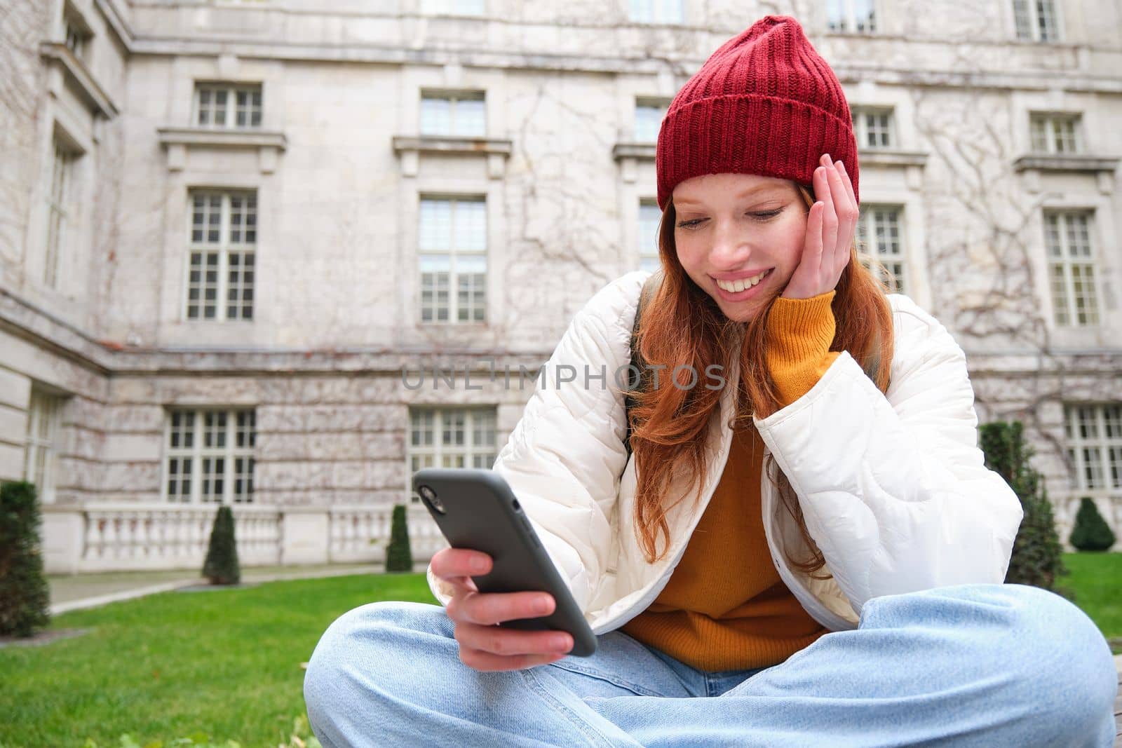 Portrait of stylish young woman, 25 years, sits on bench in park and uses mobile phone, reads online news, messages or watches video on smartphone app, connects to public wifi by Benzoix