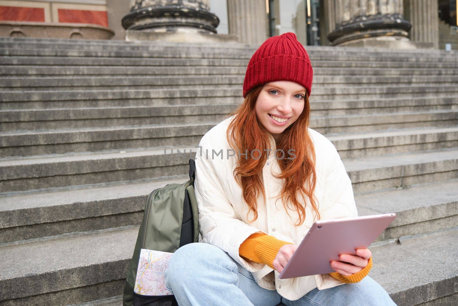 Beautiful young modern girl with red hair, holds digital tablet, sits on stairs near museum and connects public internet, sends message on gadget app by Benzoix