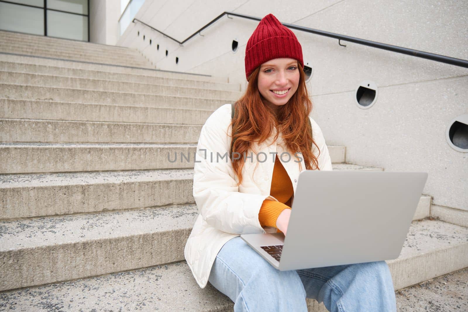 Smiling redhead girl, young woman typing on laptop keyboard, sitting outdoors on stairs with computer, working remote, doing her homework on fresh air by Benzoix