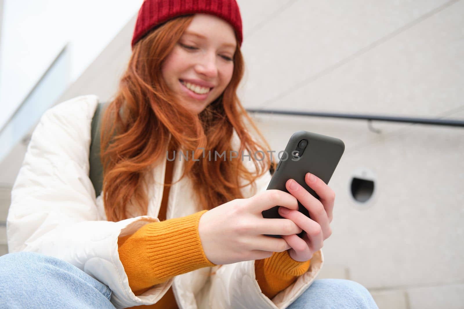 Mobile phones and people. Young stylish redhead girl sits on stairs with telephone, uses smartphone app, reads smth online by Benzoix
