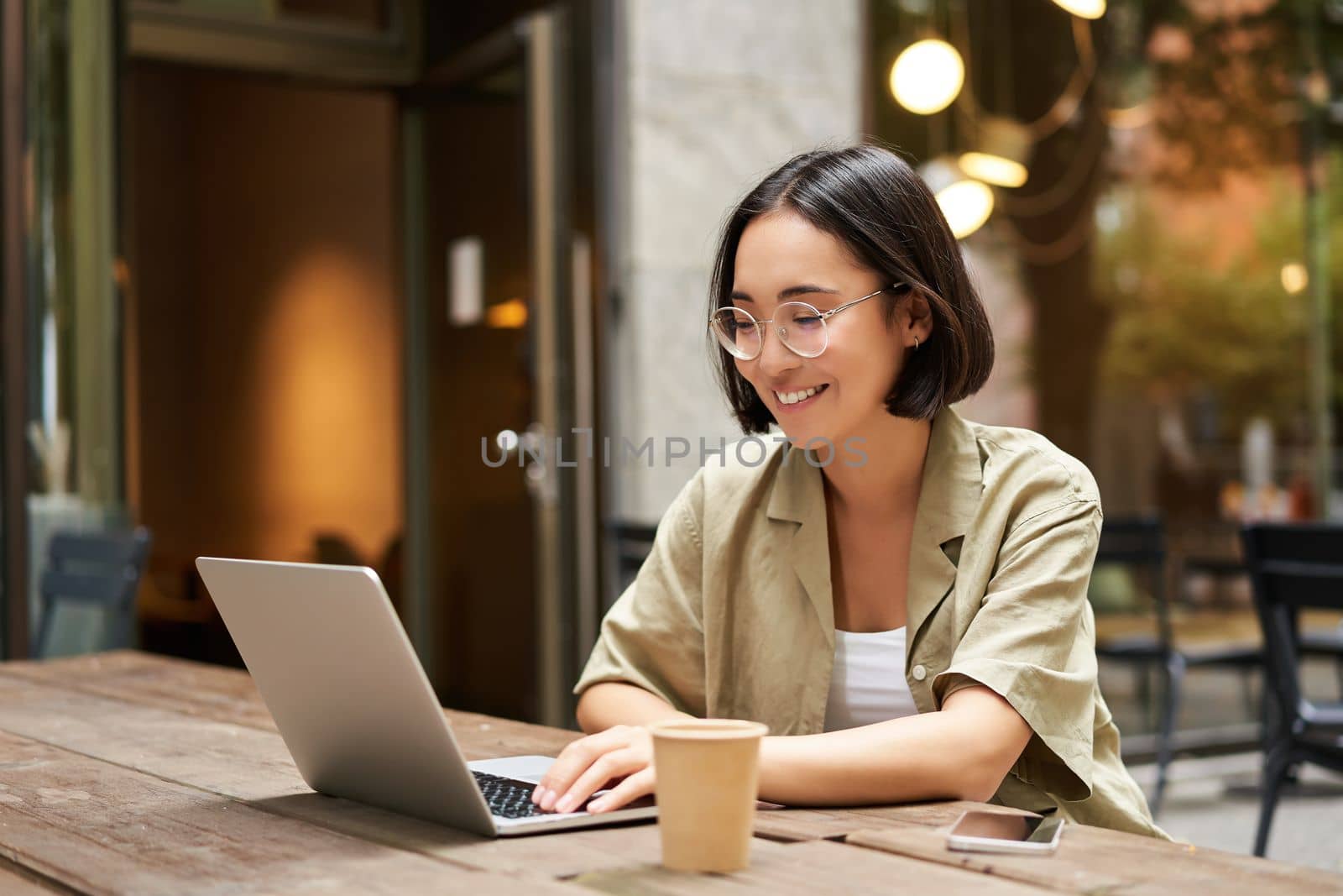 Young woman working in a cafe, using laptop and drinking coffee. Asian girl student with computer studying remotely, sitting on bench near shop.
