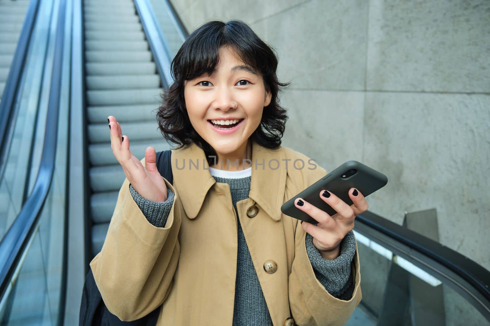 Happy triumphing asian girl, looks amazed and joyful, holds smartphone, goes down escalator with pleased face, wins, hears good news.