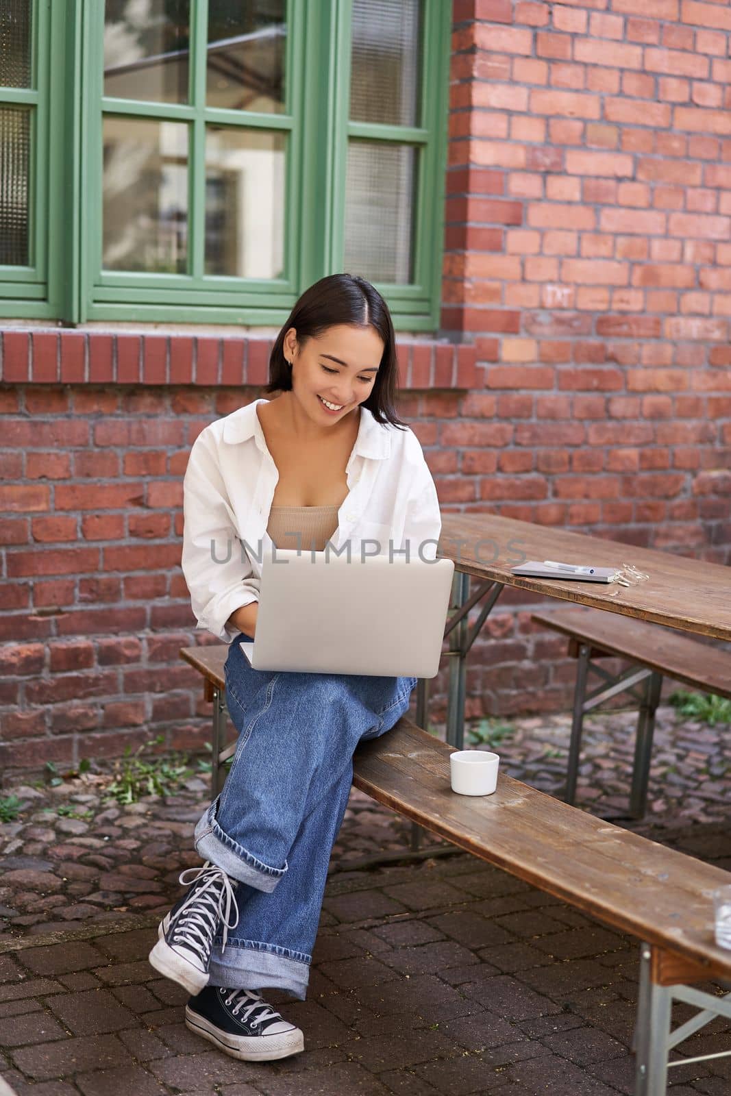 Vertical shot of asian woman working in outdoor cafe, sitting with laptop on bench and drinking coffee. Copy space