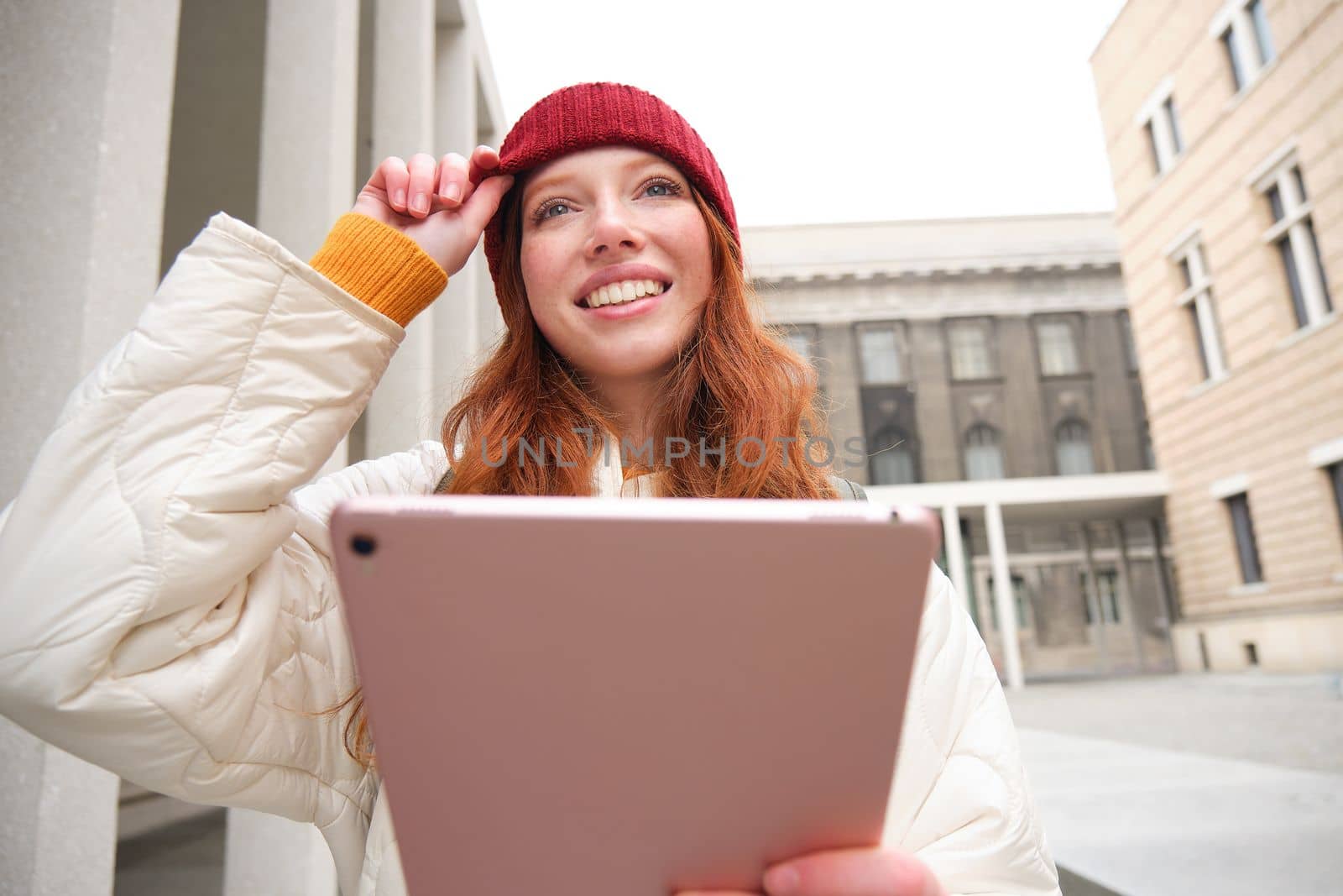Happy redhead girl in red hat, walks around city with digital tablet, connects to public internet wifi and looks for route, looks at map on her gadget.