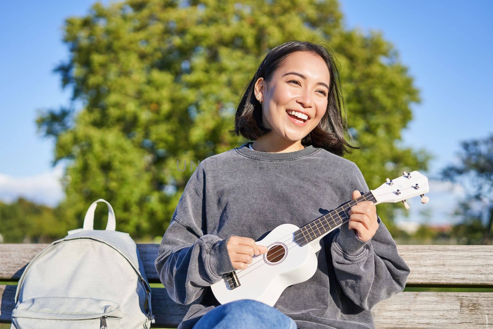 Lovely young woman sitting with backpack on bench in sunny park, plays ukulele guitar and sings along. Lifestyle