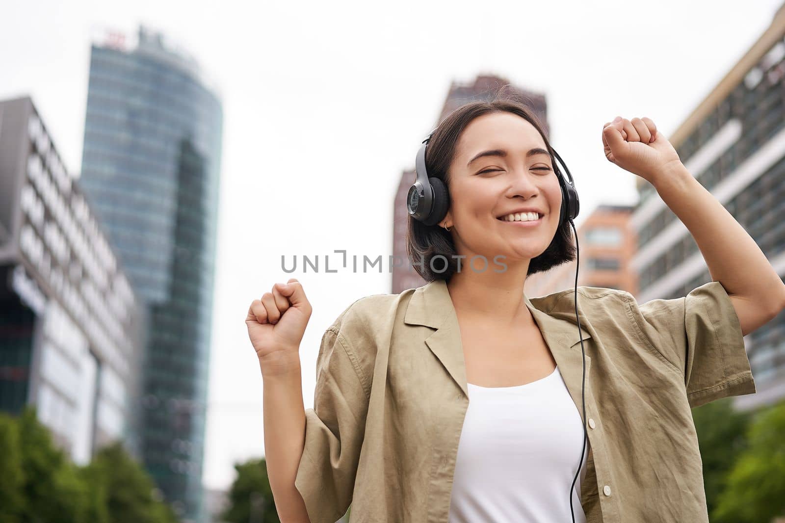 Happy asian woman in headphones, listening music and dancing on street of city centre, smiling with hands up by Benzoix