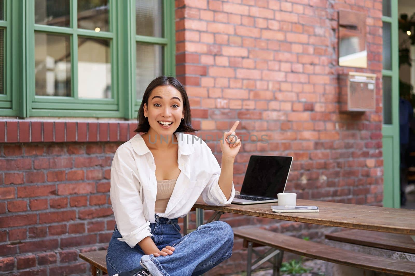 stylish modern asian girl with laptop, sitting in cafe, looking amazed and pointing at upper right corner banner, showing info advertisement by Benzoix